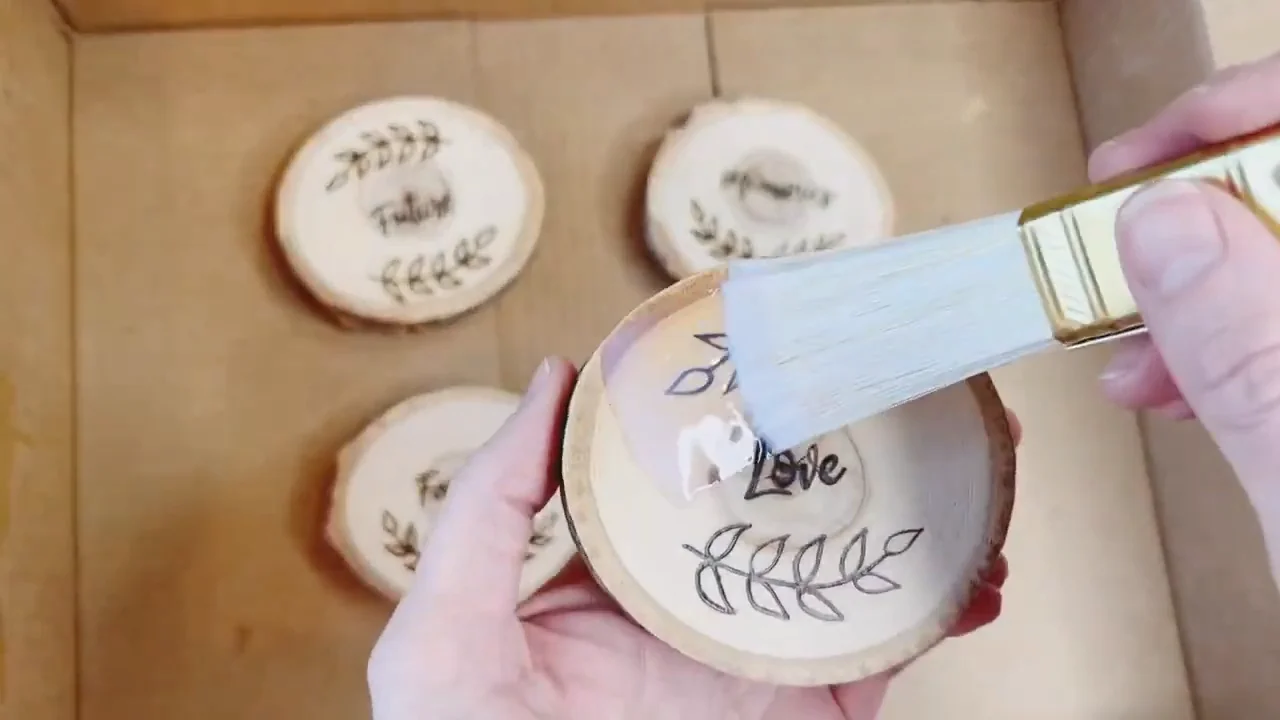 A brush applies a clear finish over a burned leaf and lettering design on a wood coaster