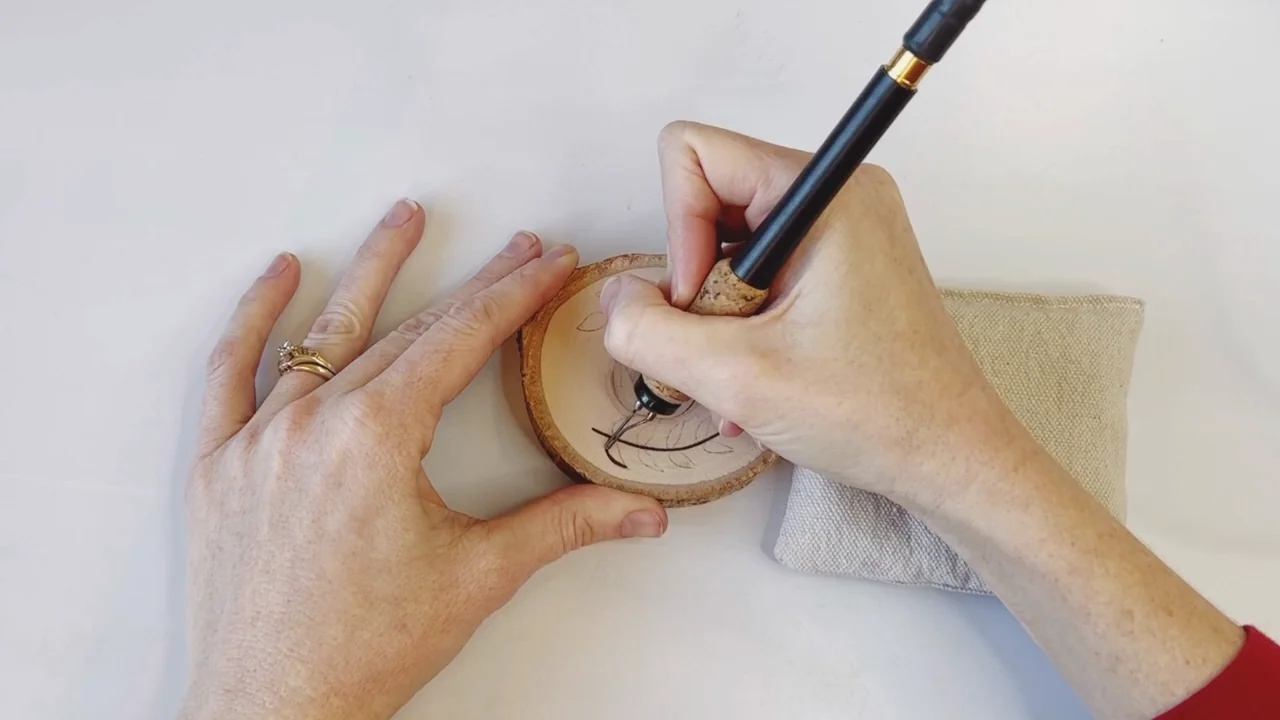 A pyrography pen follows the penciled leaf outline on a round wooden coaster