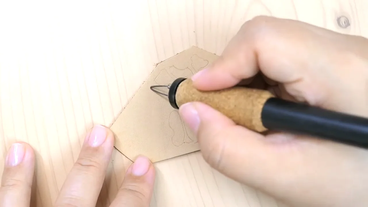 A wire-tip pyrography pen follows a light outline on a small piece of leather