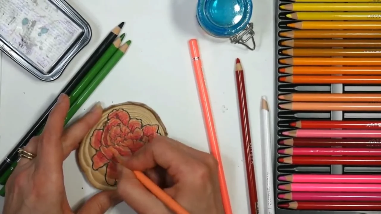 A hand adding colored-pencil layers to a flower on a wooden round