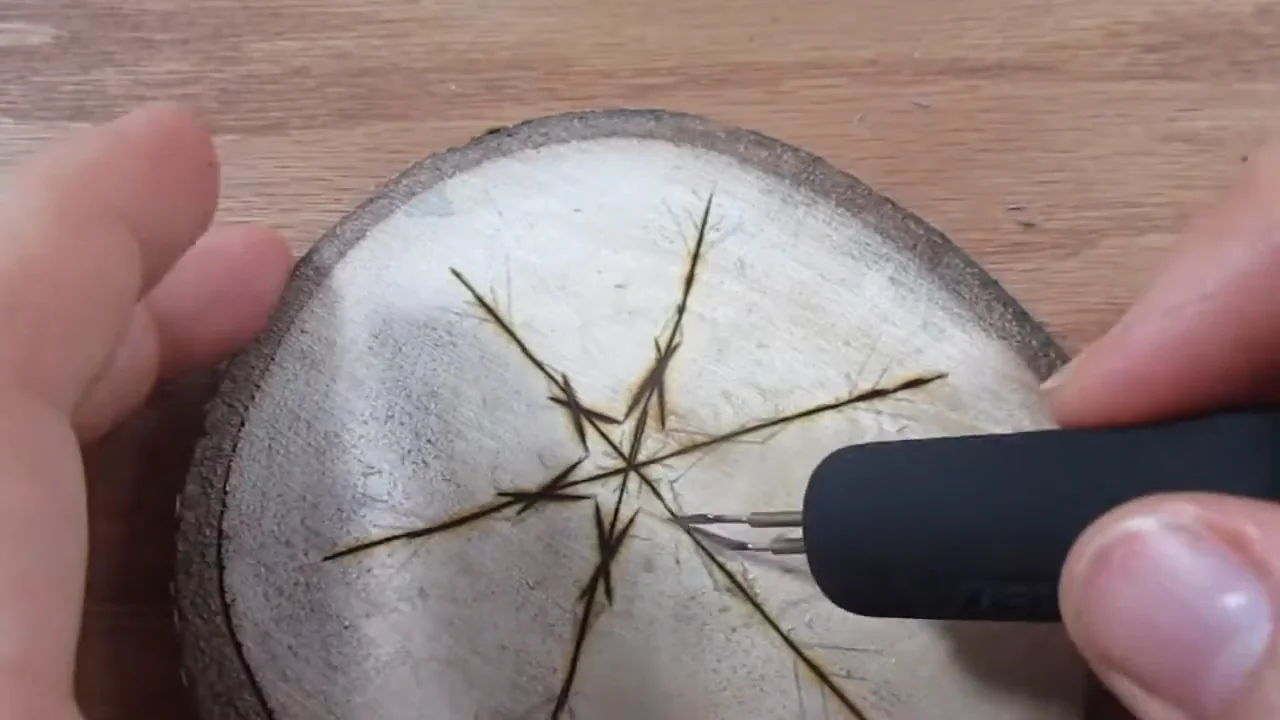 A pyrography pen burning the outline of a snowflake on a wood slice.